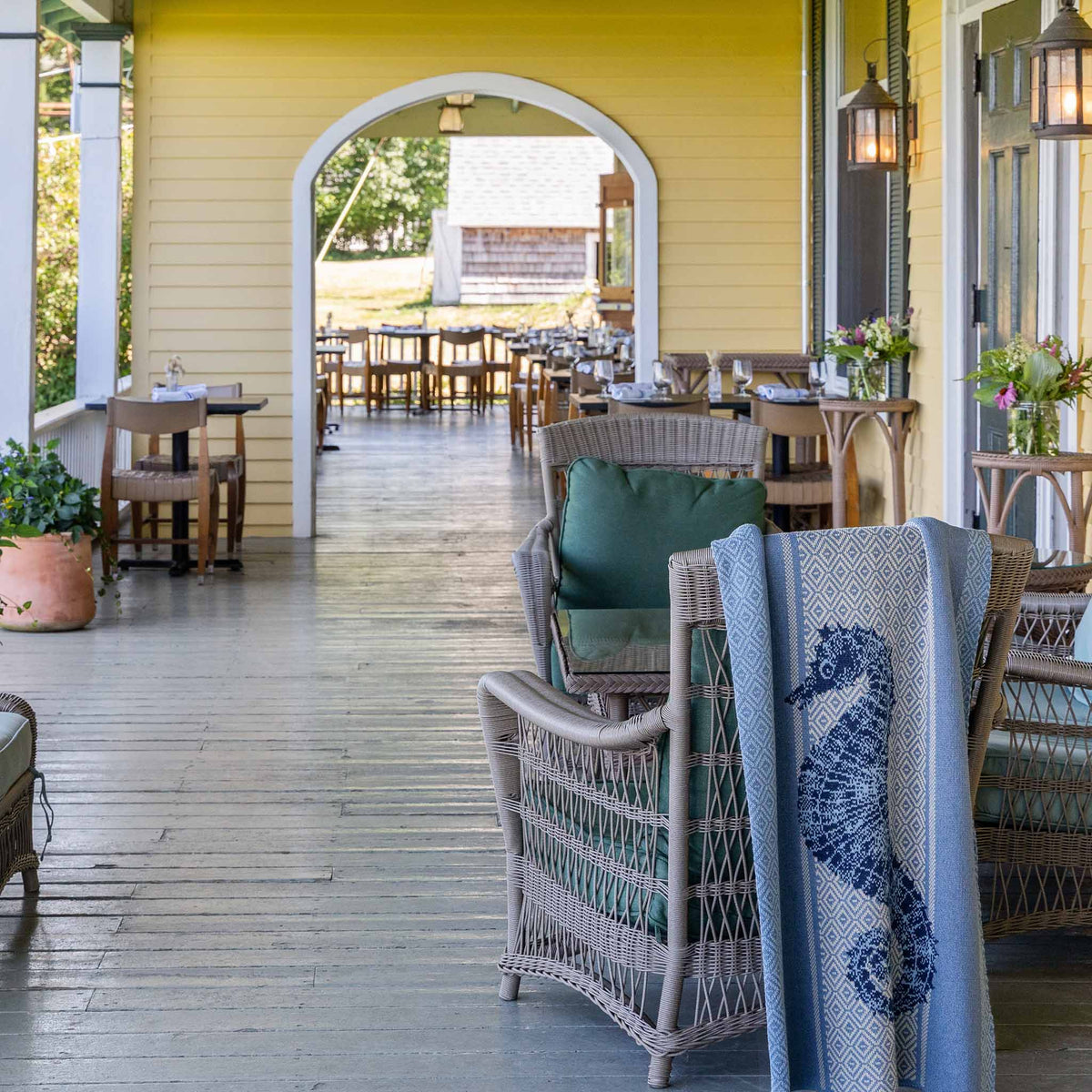 Wicker chairs and a dining table on a covered wooden porch, with the In2Green Seahorse Square - Cotton Sustainable Knit Throw Blanket in Coastal draped over one chair and an arched doorway in the background.
