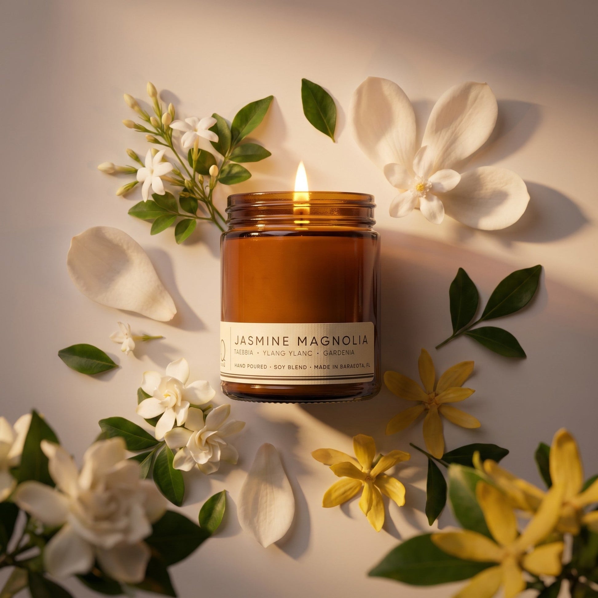 A lit amber glass candle jar labeled 'JASMINE MAGNOLIA' surrounded by white and yellow jasmine flowers and green leaves on a warm beige surface.