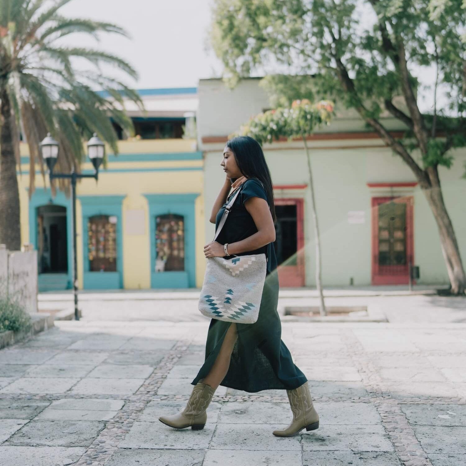 Woman carrying an AMASOUK handwoven wool Aguila Shoulder/Crossbody bag featuring a Zapotec pattern in blue, ivory, and honey on a gray backdrop. Perfect for errands, work, or casual outings.