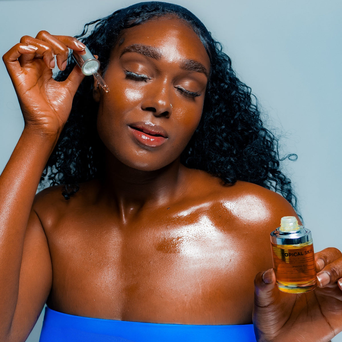 A woman with dark curly hair applies a face oil to her face while holding a bottle in her other hand, set against a solid light blue background.