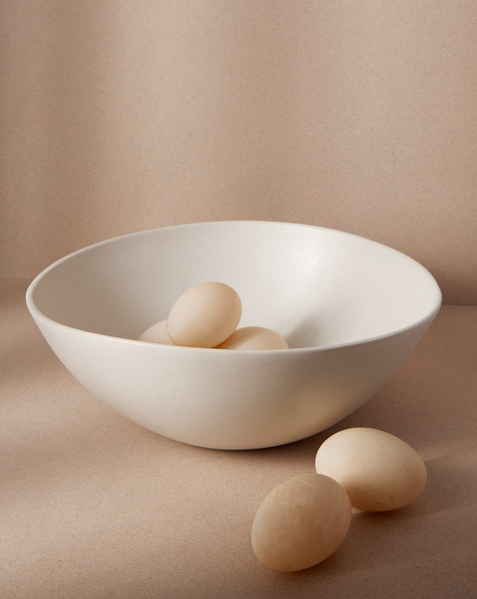 A matte white stoneware bowl with an egg inside, placed on an off-white background. Two additional eggs are positioned outside the bowl, creating a soft, minimal composition.