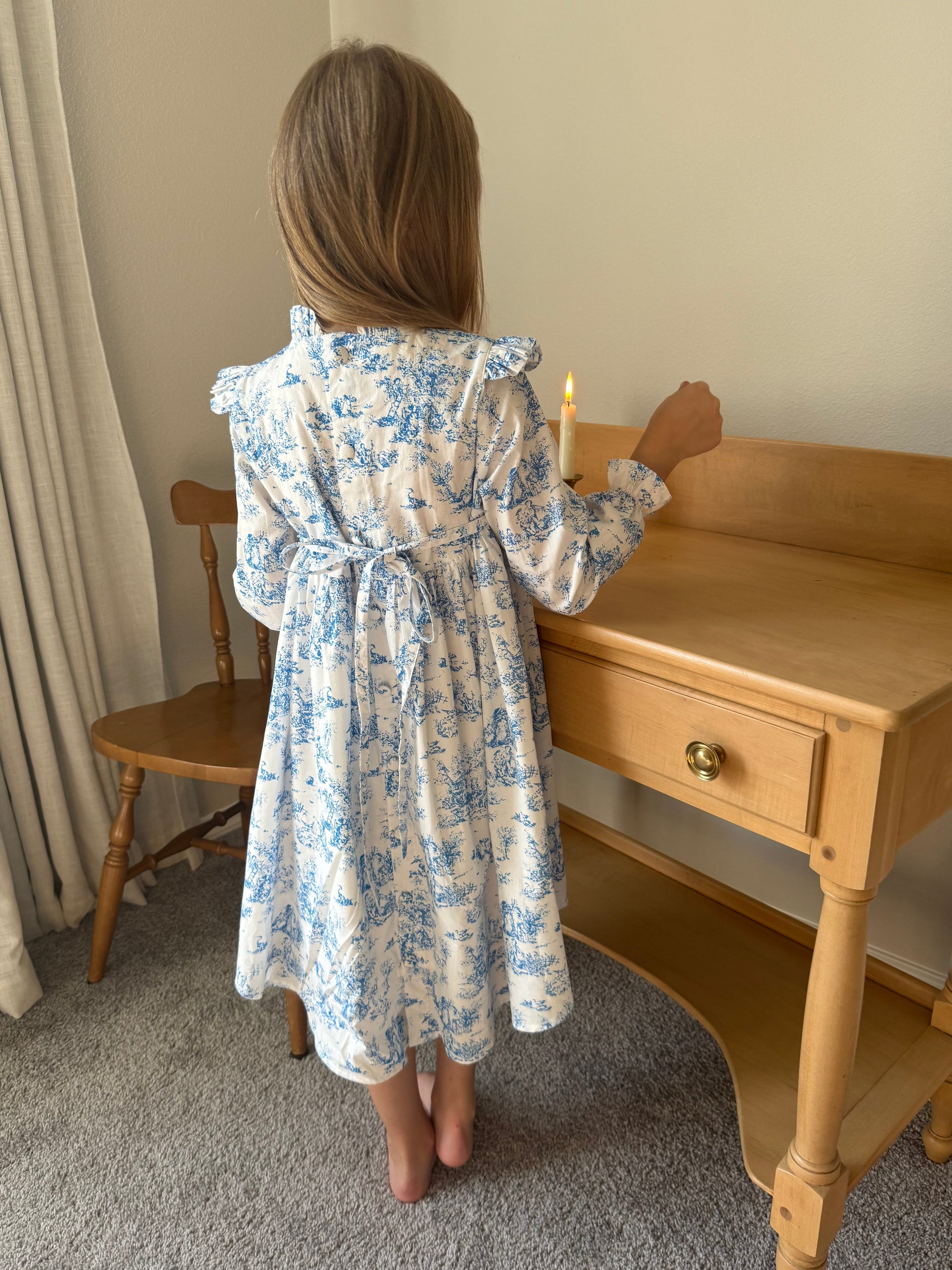 Child wearing a blue floral dress standing next to a wooden desk.