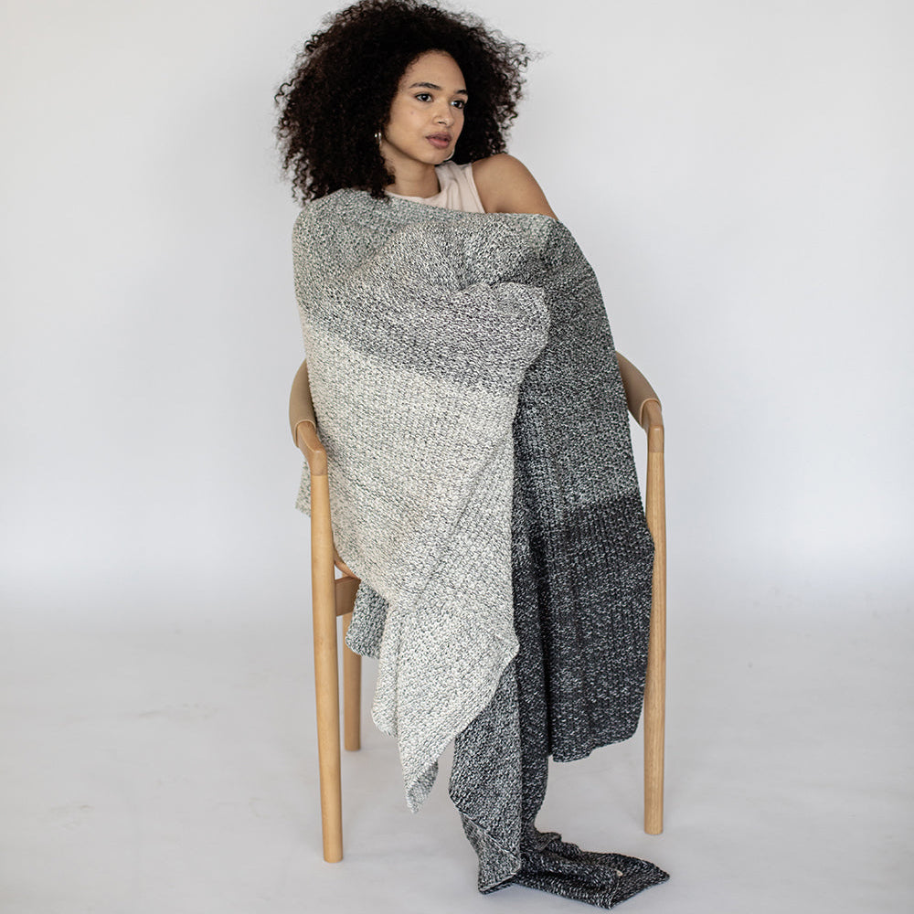 Woman sitting on a chair wrapped in a gradient gray patterned blanket against a white background.