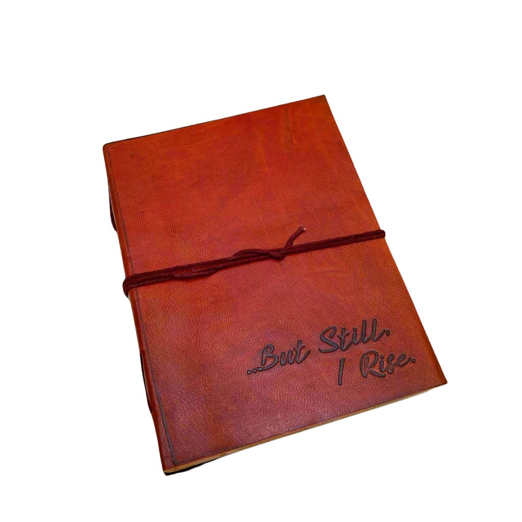 A brown leather journal with the inscription '“But Still I Rise”' embossed on the cover, tied with a red string, placed on a white wooden surface.