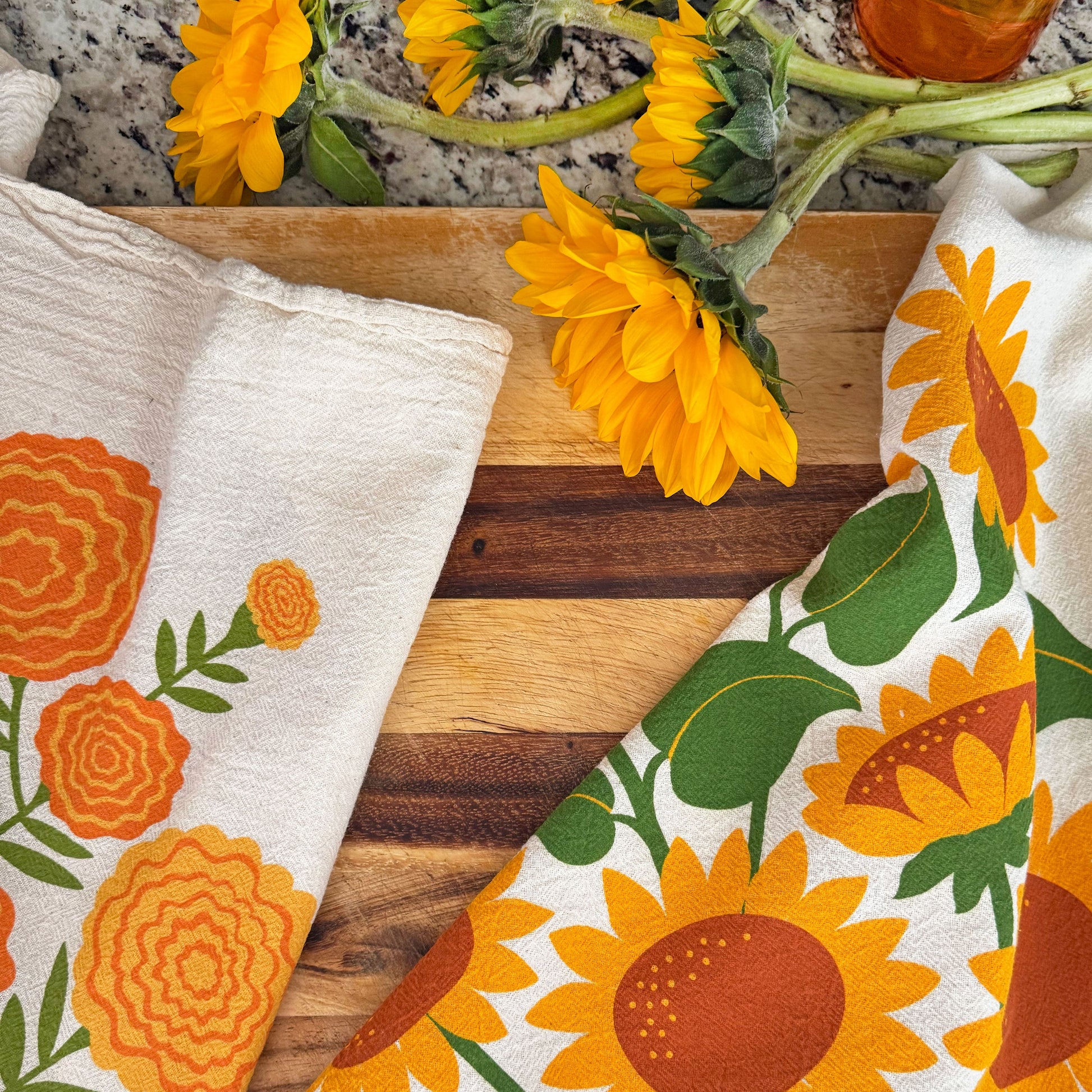 The Neighborgoods Marigold and Sunflower dish towels laid side by side on a kitchen countertop.