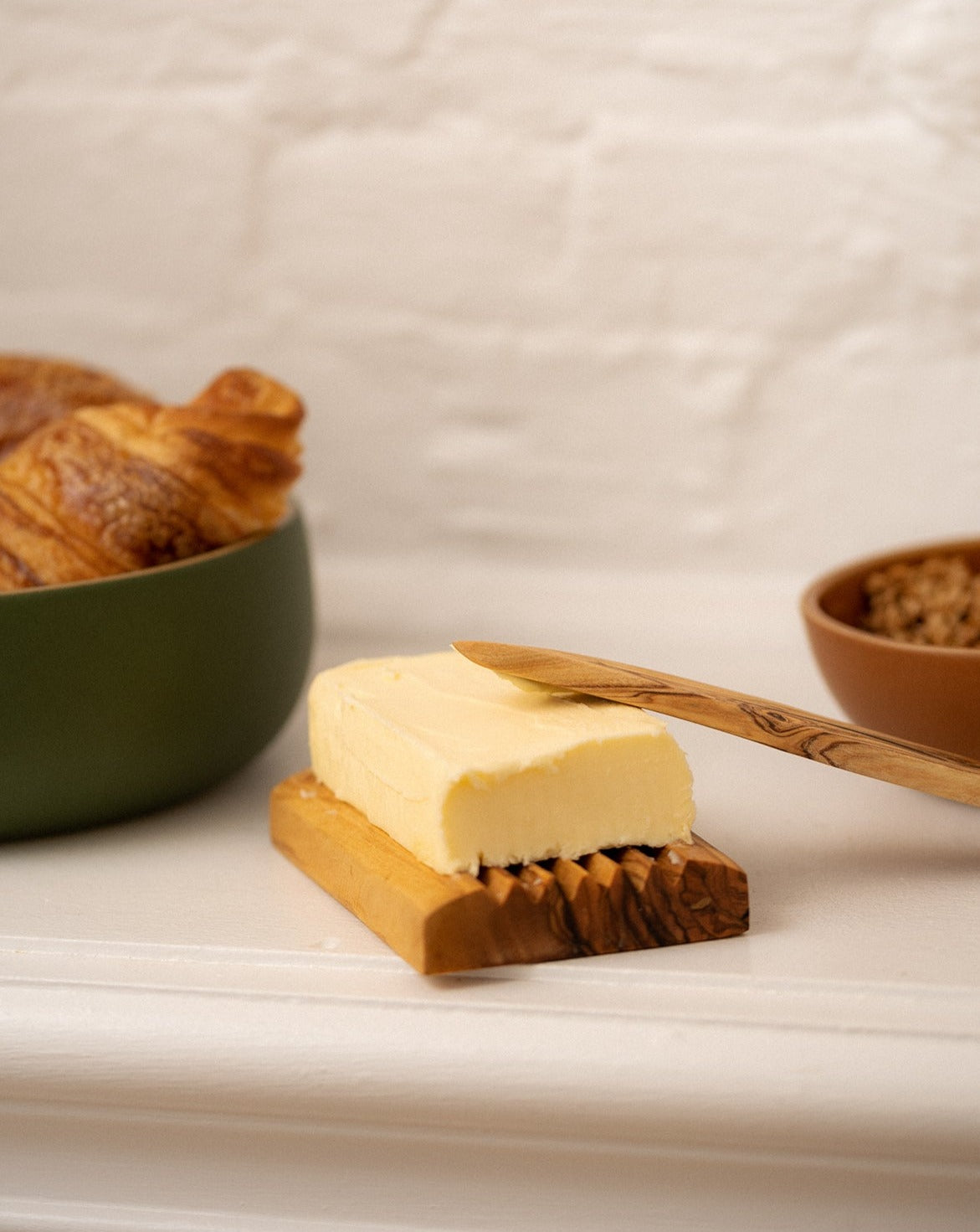 "Olive wood soap holder repurposed as a butter dish, with a wooden spreader, a bowl of bread on one side, and a smaller bowl with contents on the other side