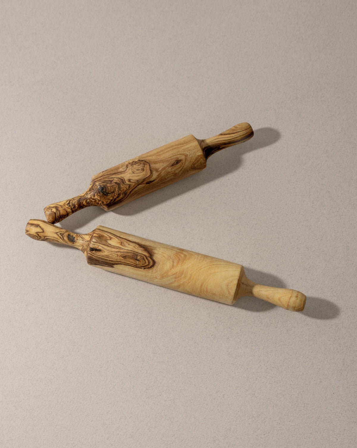 Two olivewood rolling pins, one with a flat handle and the other with a traditional cylindrical handle, lying on a grey surface.