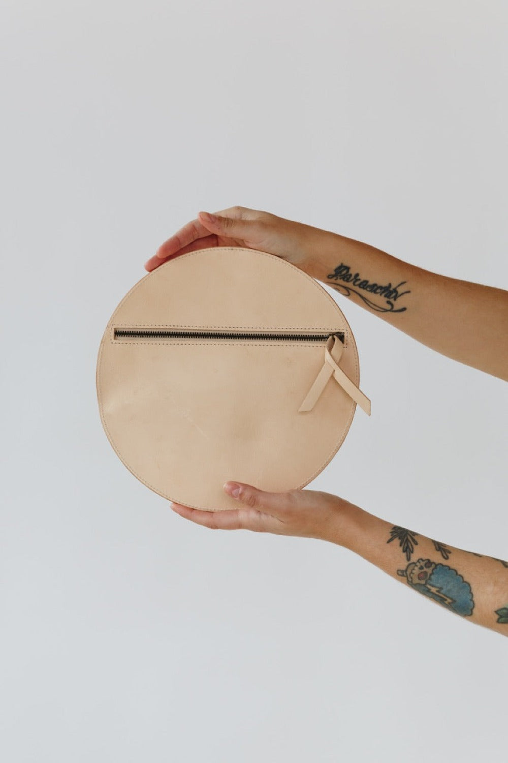 A circular natural, undyed leather clutch rests against a white background. The photo focused on the stitch and the embossed "Harper" logo.