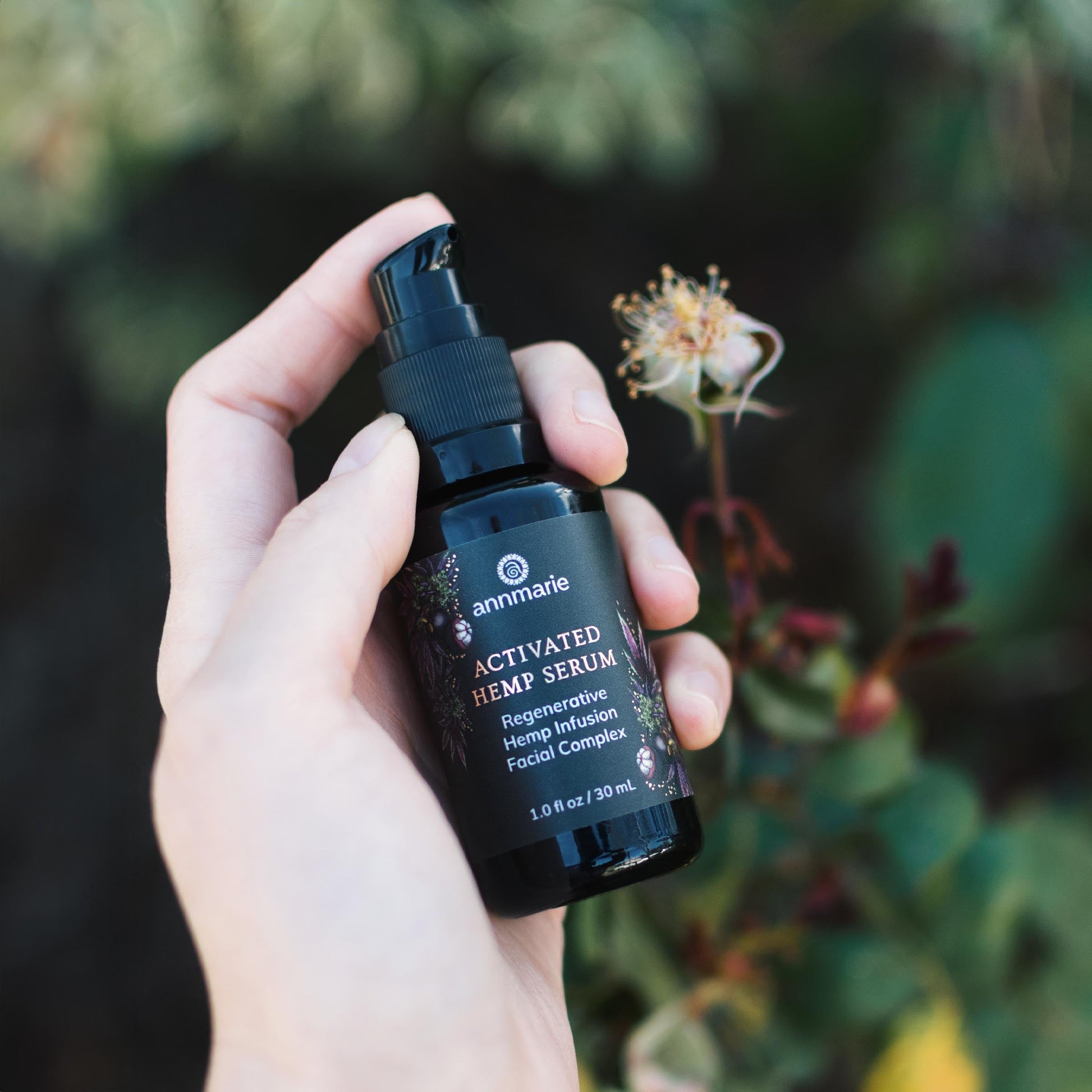 Hand holding Annmarie Activated Hemp Serum regenerative hemp infusion facial complex bottle outdoors with soft greenery and flowers in the background.
