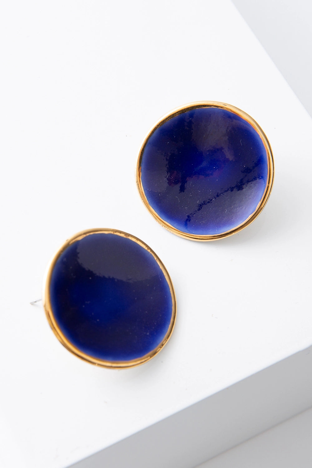 The Halo Earrings are flat, disc-shaped post earrings composed of deep blue porcelain. The discs are outlined in a ring of shining gold. The porcelain has natural variations and subtle markings, giving it a handmade look.
