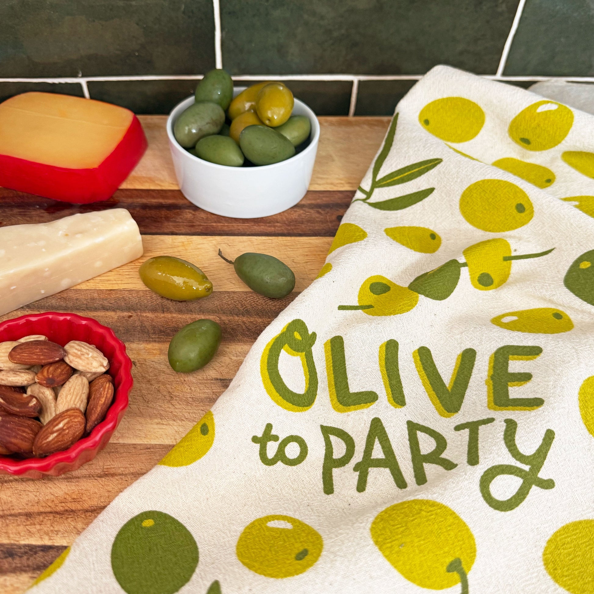 The Neighborgoods Olive dish towel featuring a olive illustration, laid next to a charcuterie display of cheese, nuts, and green olives.